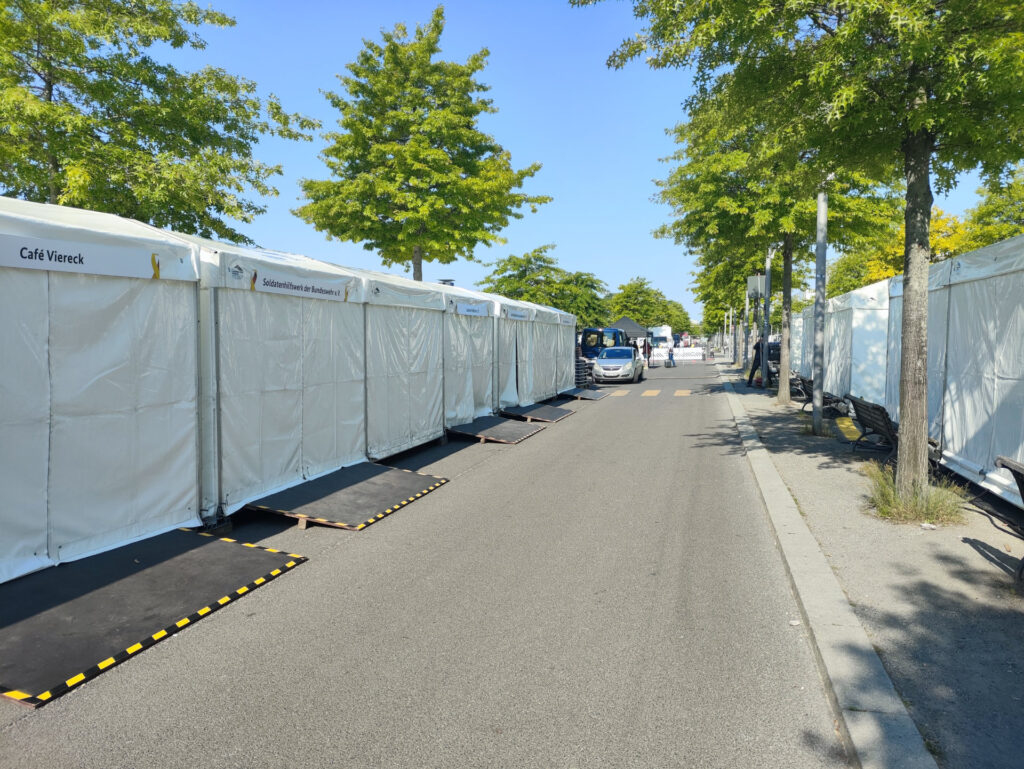 Die Kamera blickt auf eine Straße. Links und rechts stehen weiße Zelte, dazwischen stehen hier und da ein paar grüne Bäume unter dem hellblauen Himmel.