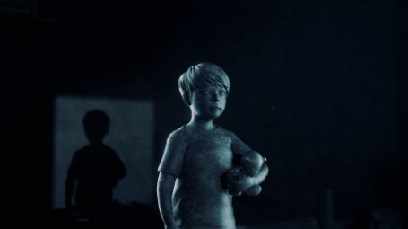 Das Bild zeigt eine Szene, die an einen Film oder ein Videospiel erinnert. Im Vordergrund steht eine junge, blonde Figur, die wie eine Statue aus Stein oder einem ähnlichen Material wirkt. Der Junge trägt ein ärmelloses Oberteil und hält ein kleines, unscharf dargestelltes Objekt, möglicherweise ein Stofftier, im Arm. Sein Gesichtsausdruck ist ernst und nachdenklich. Die Beleuchtung ist düster und kalt, mit einem bläulichen Farbton, der eine bedrückende Atmosphäre schafft. Im Hintergrund ist eine dunkle, verschwommene Silhouette einer weiteren, etwas größeren Figur zu erkennen, die wie ein Schatten oder eine unscharf dargestellte Person wirkt. Der Hintergrund selbst ist sehr dunkel und unklar, mit kaum erkennbaren Strukturen oder Objekten. Die gesamte Szene wirkt mysteriös und suggeriert eine Geschichte von Einsamkeit, Verlust oder einer unheimlichen Atmosphäre. Die Kombination aus der statuenartigen Figur des Jungen, der düsteren Beleuchtung und der undeutlichen Hintergrundsilhouette erzeugt eine beunruhigende Stimmung.